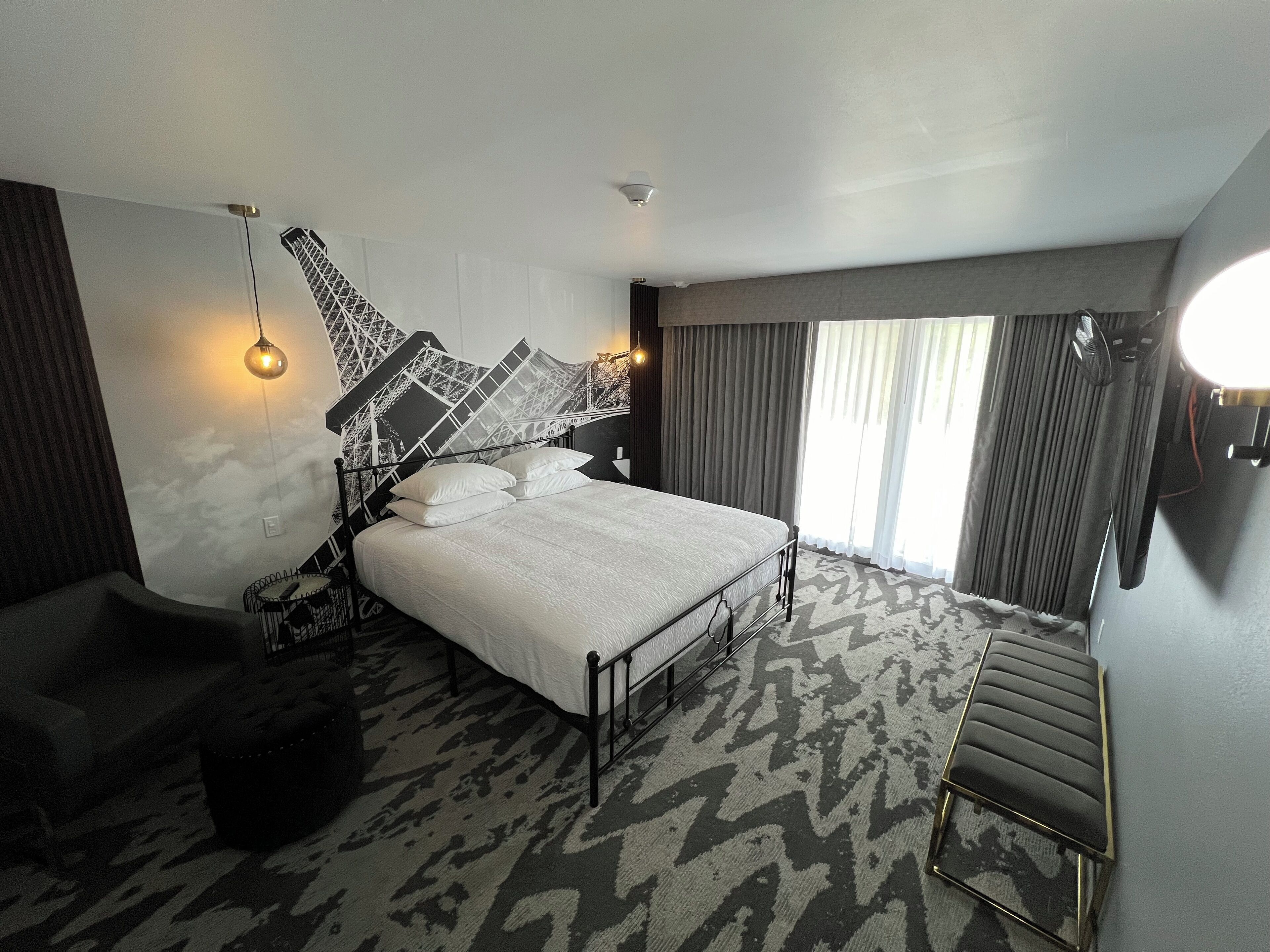 A modern hotel room with a large bed, a unique wall art piece, a bench, and a patterned carpet.