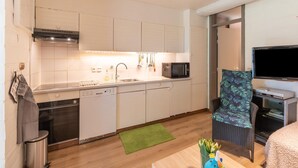 Cocina compacta con armarios blancos, microondas, lavavajillas y un lavamanos pequeño. Hay un tapete verde en el suelo y una silla de mimbre con un cojín floral.