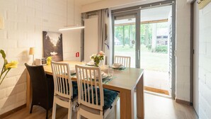 Comedor con mesa de madera, sillas blancas, un florero con flores y vistas al exterior a través de un ventanal.