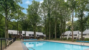 Una piscina con borde de baldosas, rodeada de árboles y un edificio con amplias ventanas.