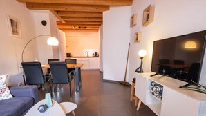 En moderne stue med sofa, spisebord, stole, et fjernsyn og et køkken i baggrunden.