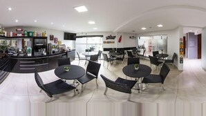 A modern cafe with round tables and black chairs, a bar area with various drinks, and a large window providing natural light. - HOLLYSTER HOTEL (Curitiba)