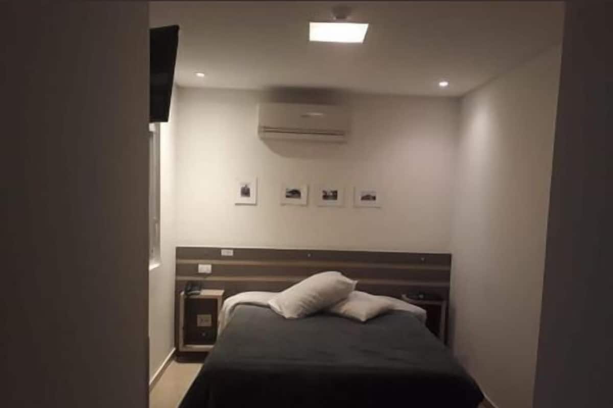 Une chambre à coucher avec un lit, une table de chevet, un téléviseur fixé au mur et un luminaire au plafond.