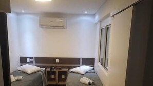 A hotel room with two single beds, a small table, a chair, an air conditioning unit, and a window. - HOLLYSTER HOTEL (Curitiba)