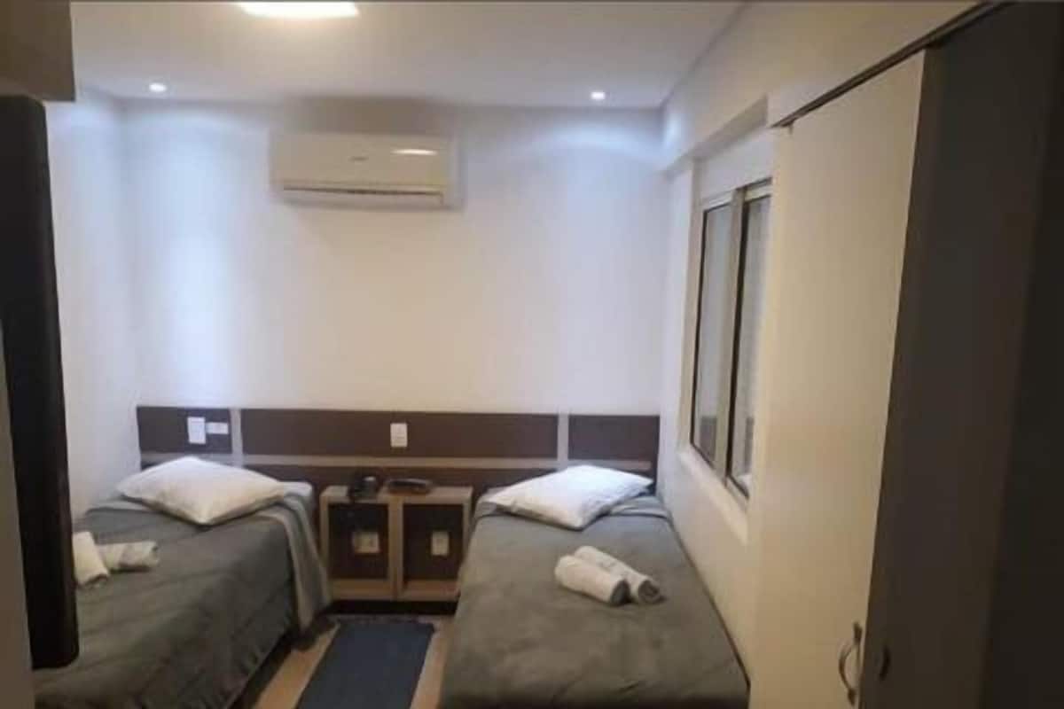 Une chambre d’hôtel avec deux lits simples, une petite table, une chaise, un appareil de climatisation et une fenêtre.