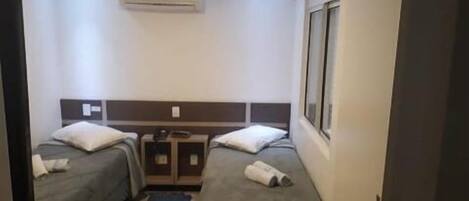 A hotel room with two single beds, a small table, a chair, an air conditioning unit, and a window.
