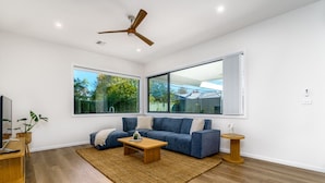 Una sala de estar moderna con un sofá azul, una mesa de centro de madera y un gran ventanal que ofrece vistas a árboles y una casa.