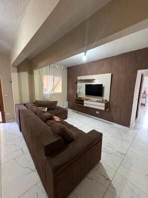 Uma sala de estar com sofá seccional, televisão de tela plana fixada na parede e janela com cortinas.