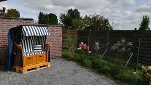 A wooden outdoor seating area with a striped canopy, a brick wall, and a garden with flowers and plants. - Charming Apartment in Norden (Norden)