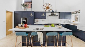 A modern kitchen with dark cabinets, a large island with bar stools, a decorative compass, and a window with a view of a brick building. - The Shippon -  Free Parking, Free WiFi, EV Charger, Coffee Machine, Large Garden (Dawlish)