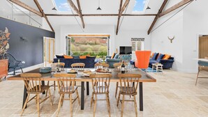 A spacious living area with a high ceiling, wooden beams, a large dining table with chairs, and a view of the outdoors through a glass door. - The Shippon -  Free Parking, Free WiFi, EV Charger, Coffee Machine, Large Garden (Dawlish)