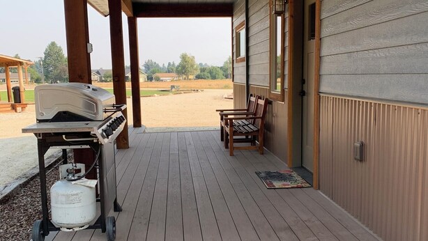 A covered porch with a grill and propane tank, wooden steps leading to a door, and a view of a grassy field. - The Guest House at Rod and Rifle Ranch (Corvallis)