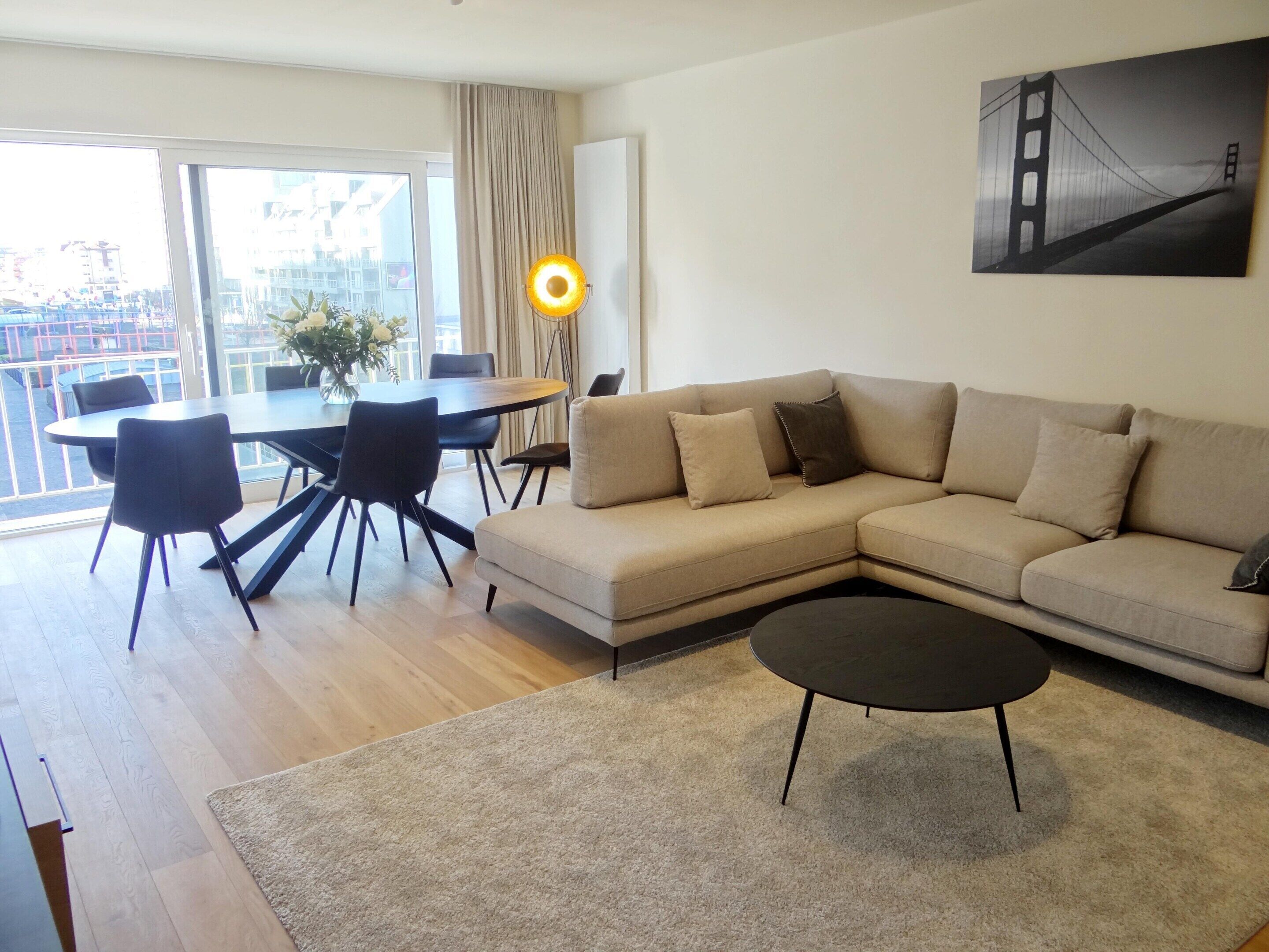 Ein modernes Wohnzimmer mit einem beigen Sofa, einem runden Esstisch mit Stühlen, einer Stehlampe und einem großen Fenster mit Blick auf Gebäude.