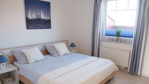 A bedroom with a bed, a nightstand, a lamp, a window with curtains, and a wall-mounted picture of sailboats. - Muschelhus Comfortable Holiday Residence (Hohwacht)