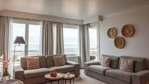 Un salon avec deux canapés, une petite table et une lampe. De grandes fenêtres, ornées de rideaux, offrent une vue sur la plage.