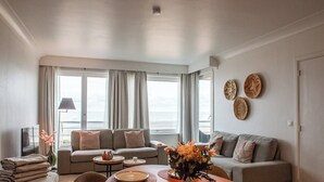 Un salon moderne avec un coin repas, des canapés gris, une table à manger en bois et une vue sur la mer grâce à de grandes fenêtres.