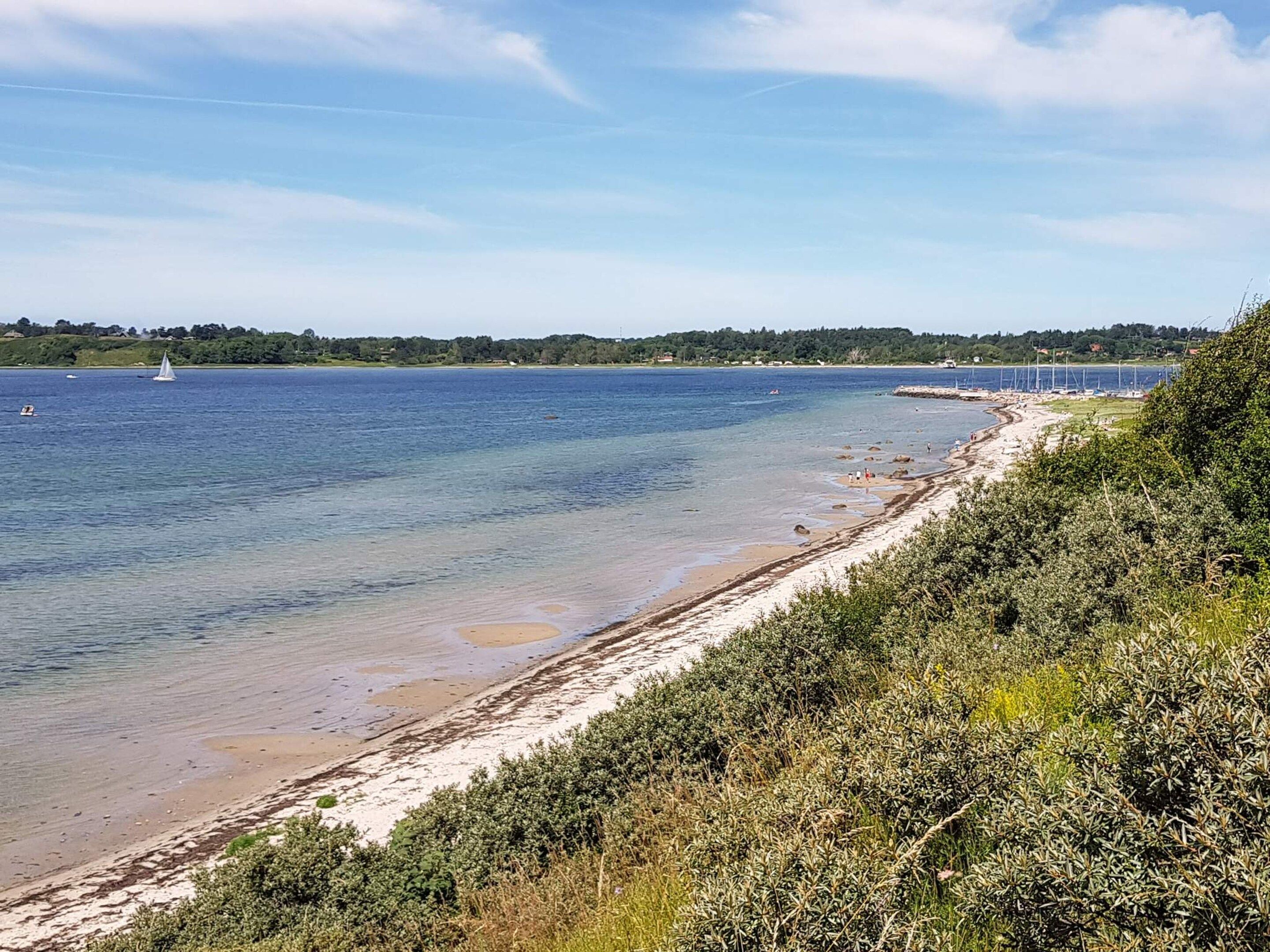 A coastal landscape with a sandy beach, clear blue water, and a distant view of a bridge.