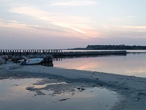 A calm waterfront scene with a pier extending into the water, a small boat docked at the pier, and a sandy beach in the foreground.