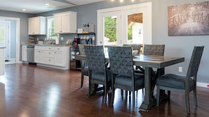 A spacious kitchen with white cabinets, a dining area with a wooden table and patterned chairs, and a large window overlooking greenery.