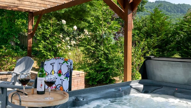 A hot tub with a wooden table and two glasses of red wine, surrounded by lush greenery and a wooden pergola.