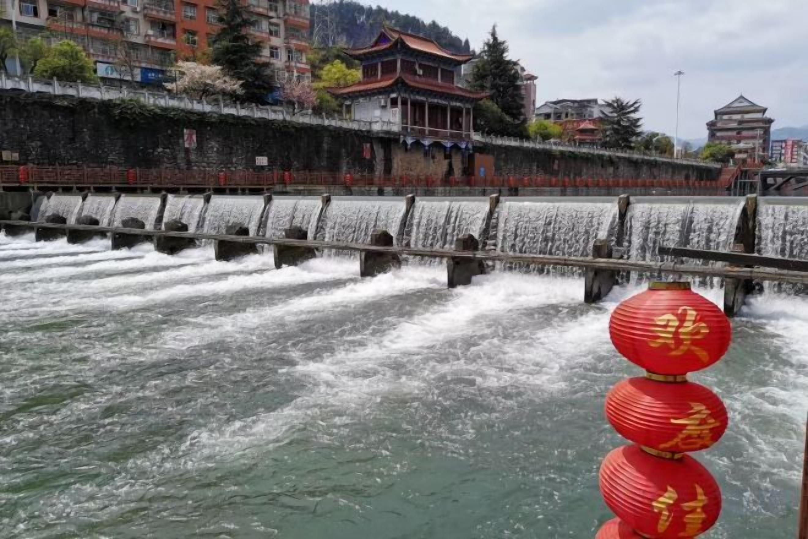A water cascade over a stone structure with traditional architecture in the background.