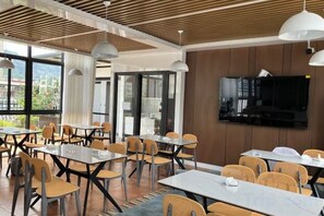 Un espace repas moderne, avec des tables et des chaises, un téléviseur à écran plat fixé au mur et de grandes fenêtres offrant une vue sur l’extérieur.