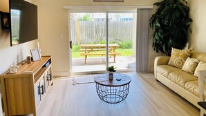 Una sala de estar con una mesa de centro de madera, un sofá, un televisor montado en la pared y un ventanal corredizo que da a un patio exterior con una mesa y sillas.