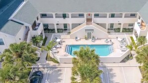 Un hotel de varios pisos con una piscina central, rodeado de palmeras y un estacionamiento.