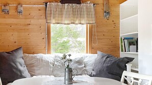 A cozy room with a wooden wall, a window with a plaid curtain, a white round table with a metal pitcher, and a bookshelf with books. - 4 Person Holiday Home in Graso (Gräsö)