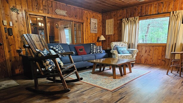 L’intérieur d’une cabane avec des murs en bois, un fauteuil à bascule, un canapé, un tapis à motifs et une fenêtre avec des rideaux.