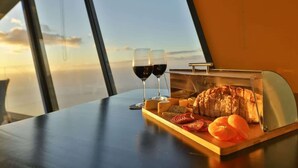 Une table avec une planche de charcuterie, deux verres de vin rouge et une vue sur le ciel par de grandes fenêtres.