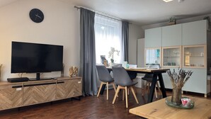 A modern living room with a flat-screen TV, wooden furniture, a dining area with chairs, and a kitchen with cabinets.