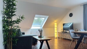 A modern living room with a sofa, a dining table, and a TV. There is a large window providing natural light and a view of the outdoors. - Kurzurlaub in Bockswiese, Hahnenklee (Goslar)
