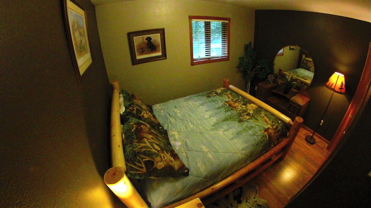 A bedroom with a bed, a bedside lamp, a window with blinds, a mirror, and a framed picture on the wall.