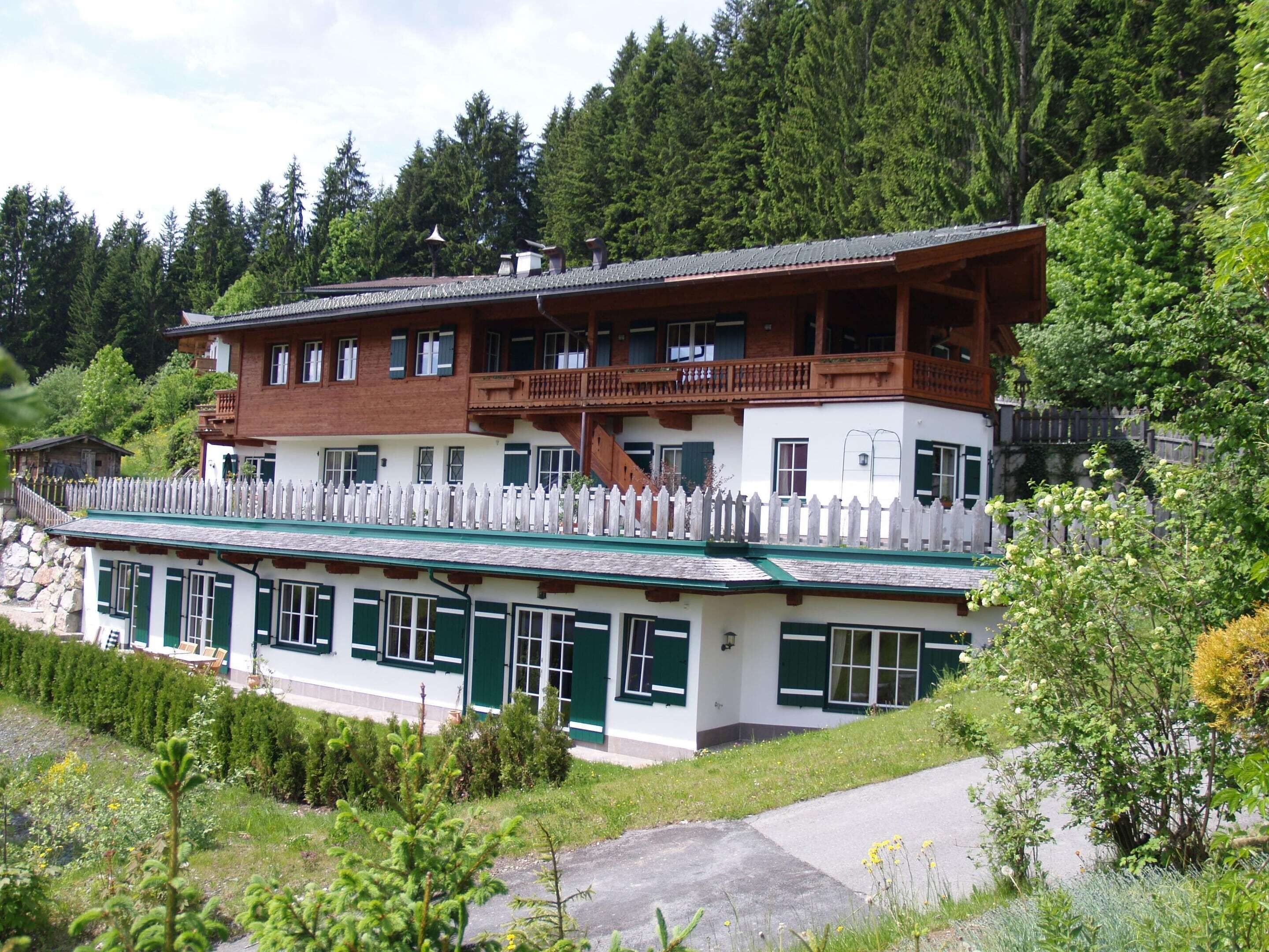 A two-story chalet with a wooden balcony, surrounded by lush greenery and a white picket fence.