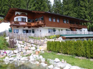 Un edificio de estilo chalé con balcón de madera, cerca blanca en forma de piquete y un jardín rocoso con un estanque.