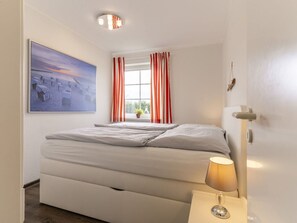 Une chambre à coucher avec un lit, une fenêtre ornée de rideaux rouges, une lampe de chevet et un tableau accroché au mur.
