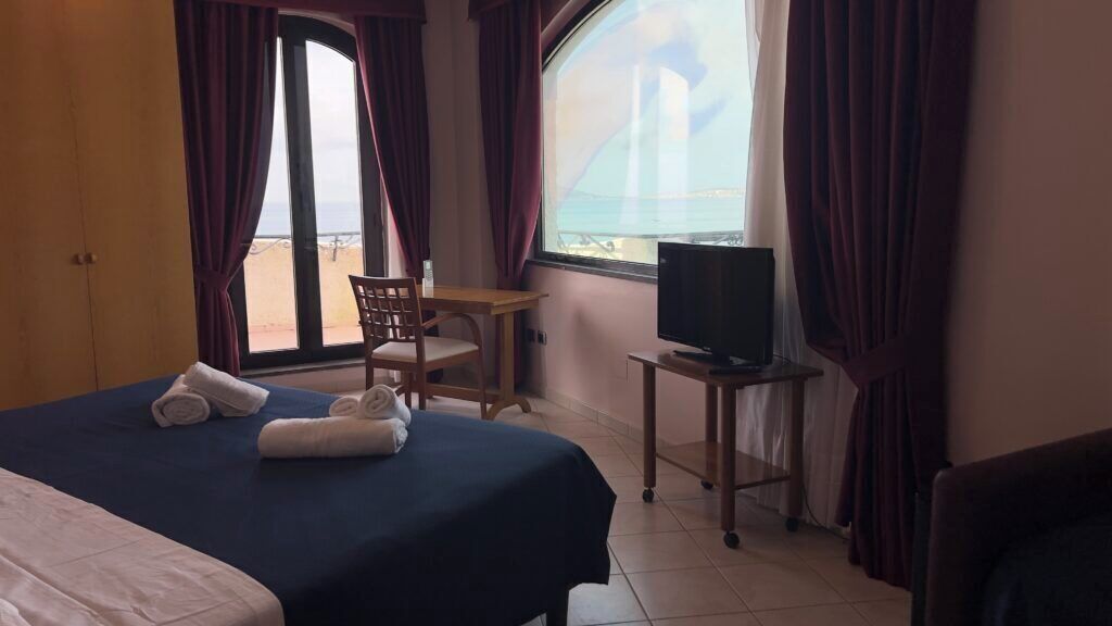 Hotellihuone, jossa on sänky, tuoli, jalustalla oleva televisio ja suuri ikkuna, josta avautuu näkymä merelle.