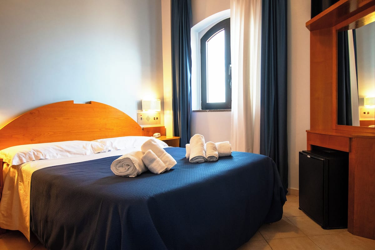 Et hotelværelse med en seng, et lille køleskab, et natbord med lampe og et vindue med gardiner.