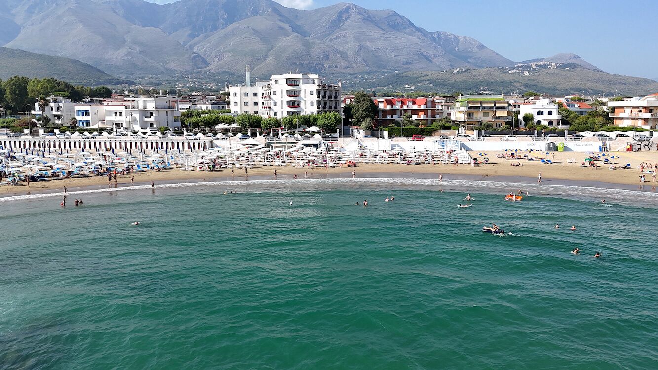 Une plage où l’on voit des gens nager et bronzer, des bâtiments en arrière-plan et des montagnes au loin.