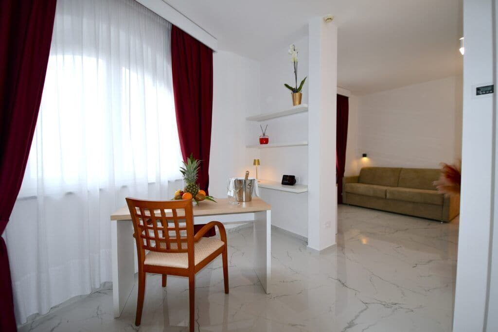 A room with a dining table, a chair, a sofa, a small shelf with decorative items, and a potted plant.