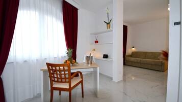 A room with a dining table, a chair, a sofa, a small shelf with decorative items, and a potted plant.