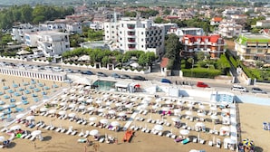 Un hotel frente a la playa con varios pisos, una piscina y numerosas sombrillas.