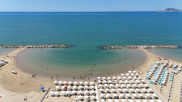 Una playa con hileras de sombrillas y sillas de descanso, un rompeolas rocoso y aguas tranquilas.