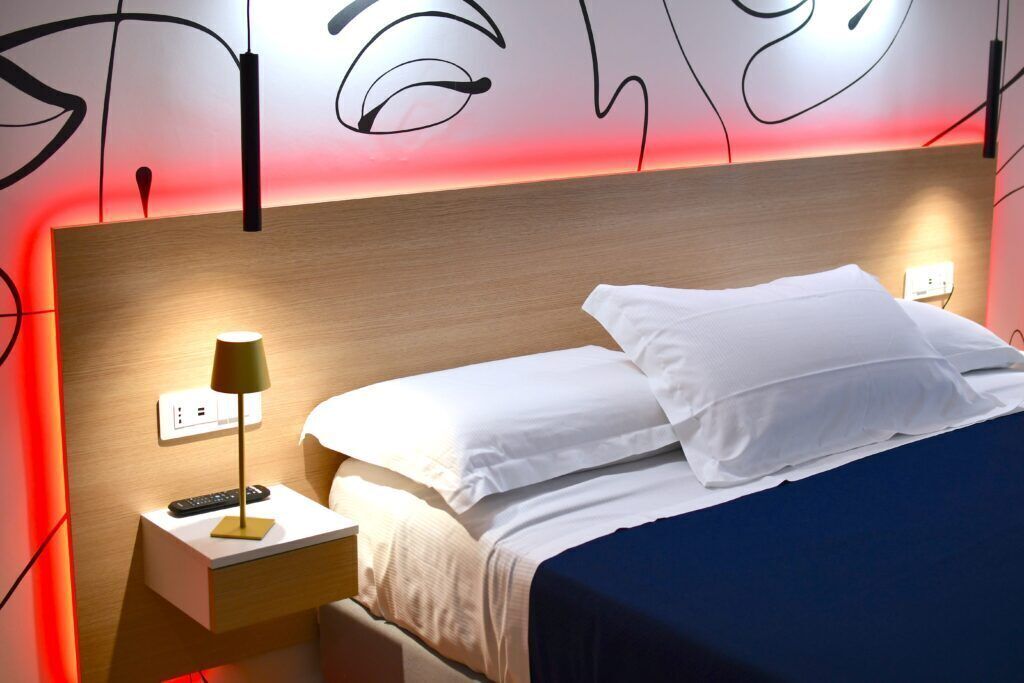 A neatly made bed with white linens, a bedside lamp, and a remote control on a wooden nightstand.