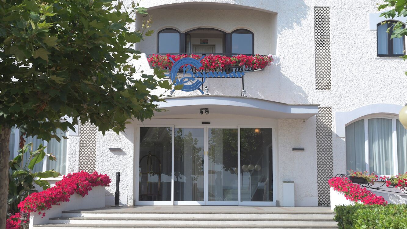 A hotel entrance with a large glass door, a balcony with red flowers, and a sign above the door.