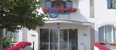 A hotel entrance with a large glass door, a balcony with red flowers, and a sign above the door.