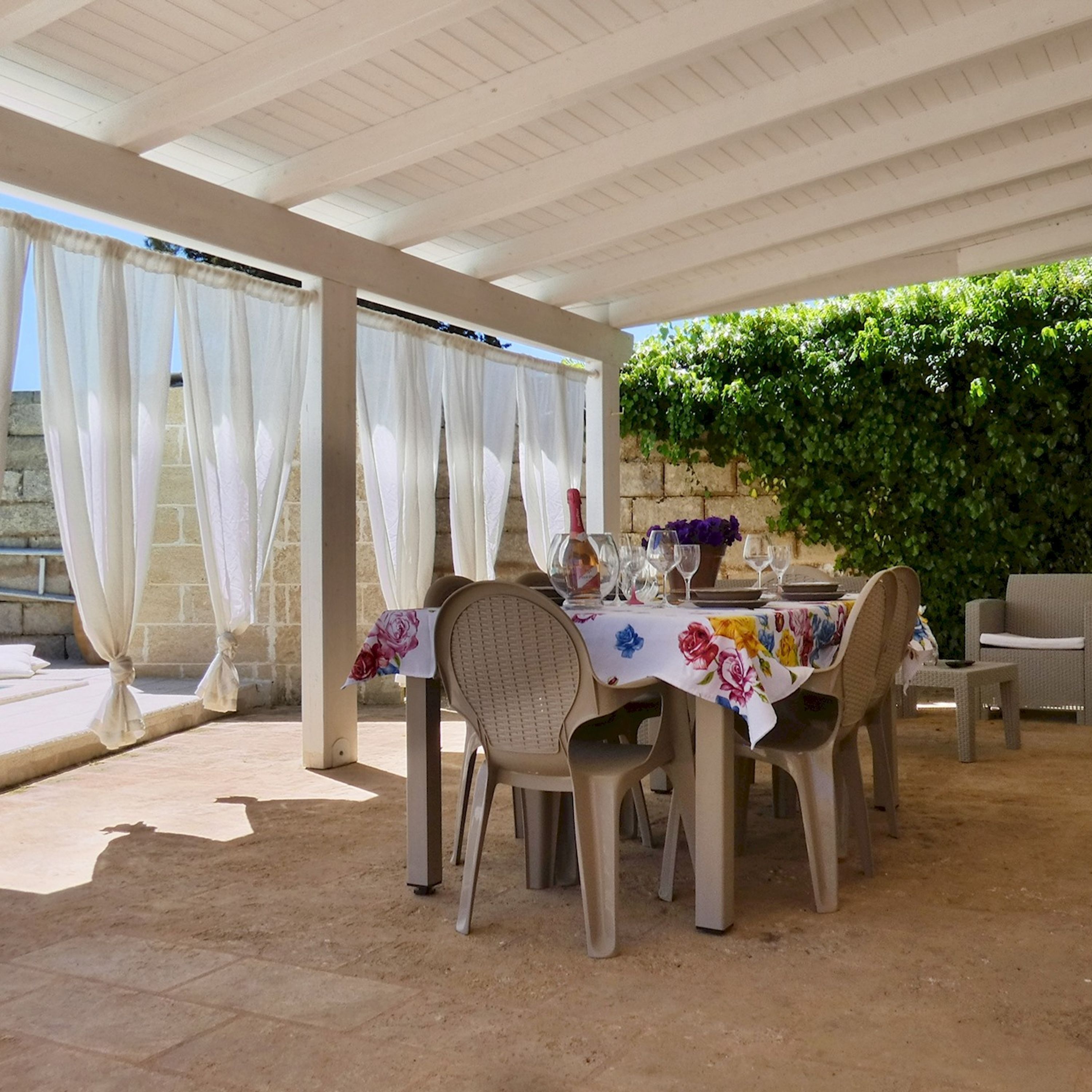 Un patio al aire libre con una mesa lista para comer, rodeada de sillas, y una zona cubierta con vistas a un muro de piedra y vegetación.