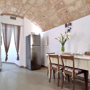 A compact kitchen with a stainless steel refrigerator, a dining table with chairs, and a vase with flowers on the wall. - La Vigna Salentina - Con Giardino e Piscina Privata (Alessano)