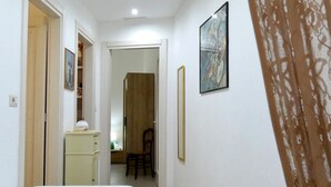 Un couloir étroit, avec un lit sur le côté gauche, une chaise au milieu et une porte au fond. Deux tableaux encadrés ornent les murs.
